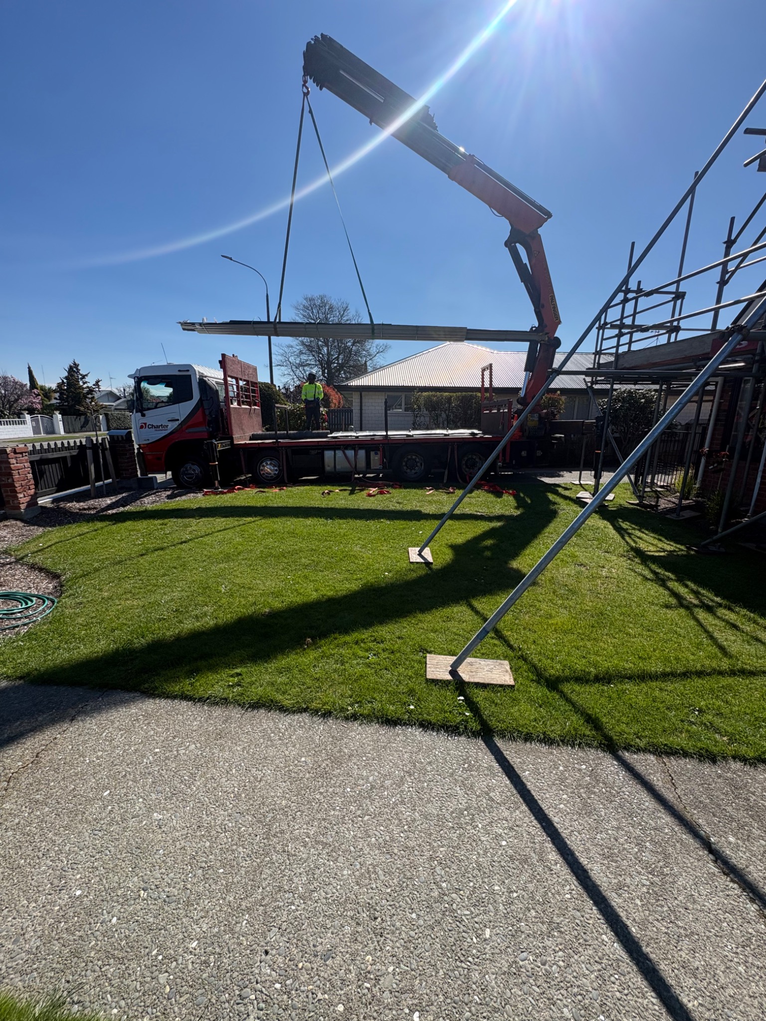 Crane lifting longrun roofing sheets from a delivery truck beside a scaffolded home